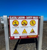 A safety sign at a glacial lagoon site with warnings depicted through symbols and text. The sign displays several cautionary messages such as not stepping on icebergs, no swimming due to freezing water, and warnings about unsafe ice for walking, dangerous currents, and rolling icebergs forming waves. Each warning is accompanied by a pictogram with a red slash, indicating prohibited actions. The sign also details emergency contact information.