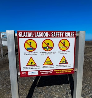 A safety sign at a glacial lagoon site with warnings depicted through symbols and text. The sign displays several cautionary messages such as not stepping on icebergs, no swimming due to freezing water, and warnings about unsafe ice for walking, dangerous currents, and rolling icebergs forming waves. Each warning is accompanied by a pictogram with a red slash, indicating prohibited actions. The sign also details emergency contact information.