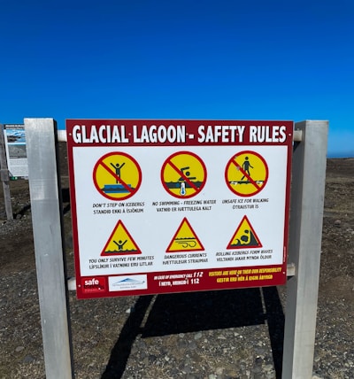 A safety sign at a glacial lagoon site with warnings depicted through symbols and text. The sign displays several cautionary messages such as not stepping on icebergs, no swimming due to freezing water, and warnings about unsafe ice for walking, dangerous currents, and rolling icebergs forming waves. Each warning is accompanied by a pictogram with a red slash, indicating prohibited actions. The sign also details emergency contact information.