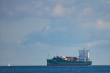 A large cargo ship is sailing across a calm body of water under a mostly cloudy sky. The vessel is fully loaded with colorful shipping containers stacked on its deck. In the background, a small sailboat is visible on the left side of the image.