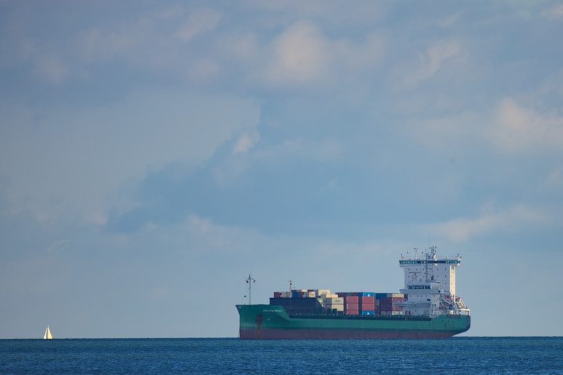 A large cargo ship is sailing across a calm body of water under a mostly cloudy sky. The vessel is fully loaded with colorful shipping containers stacked on its deck. In the background, a small sailboat is visible on the left side of the image.