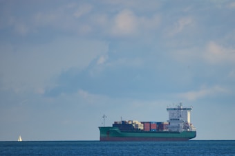 A large cargo ship is sailing across a calm body of water under a mostly cloudy sky. The vessel is fully loaded with colorful shipping containers stacked on its deck. In the background, a small sailboat is visible on the left side of the image.