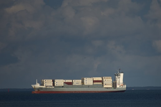 A cargo ship is sailing on a calm sea under a partly cloudy sky. The vessel is carrying a large number of shipping containers, stacked neatly on its deck, with some bearing visible logos. The ship cuts through the water, leaving a small wake behind.