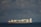 A cargo ship is sailing on a calm sea under a partly cloudy sky. The vessel is carrying a large number of shipping containers, stacked neatly on its deck, with some bearing visible logos. The ship cuts through the water, leaving a small wake behind.