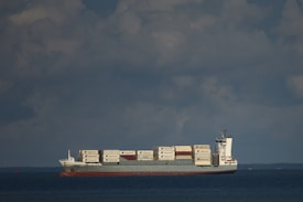 A cargo ship is sailing on a calm sea under a partly cloudy sky. The vessel is carrying a large number of shipping containers, stacked neatly on its deck, with some bearing visible logos. The ship cuts through the water, leaving a small wake behind.