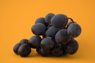 A cluster of dark purple grapes rests on a vibrant orange background, creating a strong contrast. The smooth surface of the grapes reflects light subtly, highlighting their plump and fresh appearance. The stem is partially visible, adding a natural element to the composition.