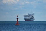 A large cargo ship filled with numerous shipping containers sails across a vast, calm ocean under a slightly cloudy sky. In the foreground, a red buoy marked with letters is visible, floating on the water.