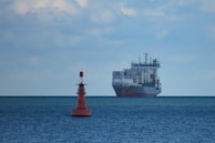 A large cargo ship filled with numerous shipping containers sails across a vast, calm ocean under a slightly cloudy sky. In the foreground, a red buoy marked with letters is visible, floating on the water.