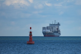 A large cargo ship filled with numerous shipping containers sails across a vast, calm ocean under a slightly cloudy sky. In the foreground, a red buoy marked with letters is visible, floating on the water.