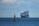A large cargo ship filled with numerous shipping containers sails across a vast, calm ocean under a slightly cloudy sky. In the foreground, a red buoy marked with letters is visible, floating on the water.