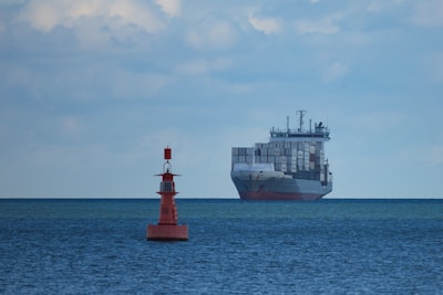 A large cargo ship filled with numerous shipping containers sails across a vast, calm ocean under a slightly cloudy sky. In the foreground, a red buoy marked with letters is visible, floating on the water.