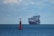 A large cargo ship filled with numerous shipping containers sails across a vast, calm ocean under a slightly cloudy sky. In the foreground, a red buoy marked with letters is visible, floating on the water.