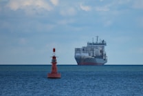 A large cargo ship filled with numerous shipping containers sails across a vast, calm ocean under a slightly cloudy sky. In the foreground, a red buoy marked with letters is visible, floating on the water.