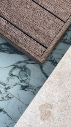 A section of wooden deck planks next to an area featuring marble-like tiles and a light-colored stone surface. The textures of the materials are distinct, with the wooden planks showing a rough grain, while the tiles have smooth, swirling dark patterns on a white background.