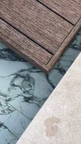 A section of wooden deck planks next to an area featuring marble-like tiles and a light-colored stone surface. The textures of the materials are distinct, with the wooden planks showing a rough grain, while the tiles have smooth, swirling dark patterns on a white background.
