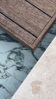 A section of wooden deck planks next to an area featuring marble-like tiles and a light-colored stone surface. The textures of the materials are distinct, with the wooden planks showing a rough grain, while the tiles have smooth, swirling dark patterns on a white background.