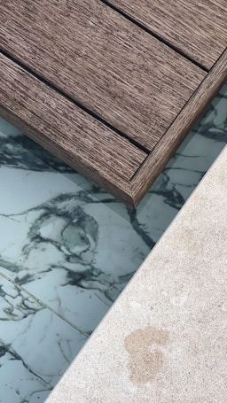 A section of wooden deck planks next to an area featuring marble-like tiles and a light-colored stone surface. The textures of the materials are distinct, with the wooden planks showing a rough grain, while the tiles have smooth, swirling dark patterns on a white background.