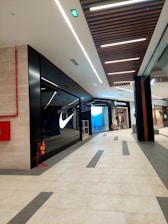 The image depicts the interior of a modern shopping mall. There is a well-known sportswear store with a prominent illuminated logo on the left side. Opposite to it, there is another store which appears to deal in clothing. The floor is tiled with a combination of light and dark tiles in a geometric pattern. The ceiling features linear lighting and wooden panels. A red fire extinguisher is mounted on the wall on the left.
