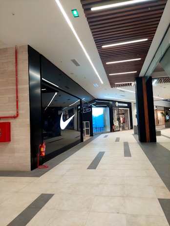 The image depicts the interior of a modern shopping mall. There is a well-known sportswear store with a prominent illuminated logo on the left side. Opposite to it, there is another store which appears to deal in clothing. The floor is tiled with a combination of light and dark tiles in a geometric pattern. The ceiling features linear lighting and wooden panels. A red fire extinguisher is mounted on the wall on the left.