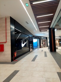 The image depicts the interior of a modern shopping mall. There is a well-known sportswear store with a prominent illuminated logo on the left side. Opposite to it, there is another store which appears to deal in clothing. The floor is tiled with a combination of light and dark tiles in a geometric pattern. The ceiling features linear lighting and wooden panels. A red fire extinguisher is mounted on the wall on the left.