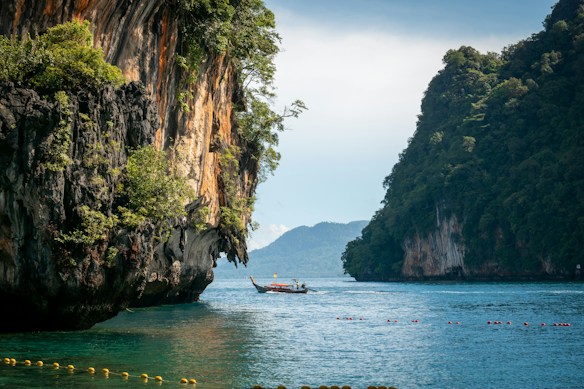 A serene seascape with towering limestone cliffs covered in lush greenery on either side of a calm, blue body of water. A traditional long-tail boat is floating in the middle of the water, with distant mountains visible in the background under a clear sky.