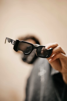 A hand is holding a pair of black, oversized sunglasses with a blurred background. The focus is on the sunglasses' frame, with light reflections visible on the lenses.