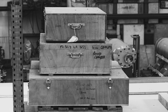 A stack of three wooden crates, each with metal latches and writing on the front. The crates are arranged from largest at the bottom to smallest on top, placed in an industrial setting with machinery and pipes in the background. The scene is captured in black and white, emphasizing textures and shapes.