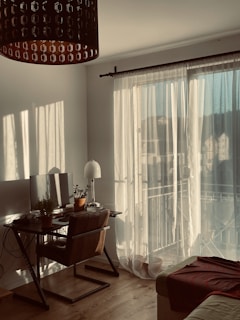A cozy room filled with warm sunlight filtering through sheer curtains. A wooden desk is adorned with a computer monitor and several potted plants. A comfortable chair is positioned at the desk, and a floor lamp stands nearby. A soft, brown blanket is draped over the edge of a sofa. The wooden floor adds to the inviting atmosphere.
