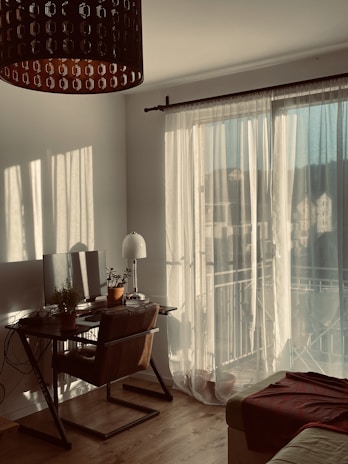 A cozy room filled with warm sunlight filtering through sheer curtains. A wooden desk is adorned with a computer monitor and several potted plants. A comfortable chair is positioned at the desk, and a floor lamp stands nearby. A soft, brown blanket is draped over the edge of a sofa. The wooden floor adds to the inviting atmosphere.
