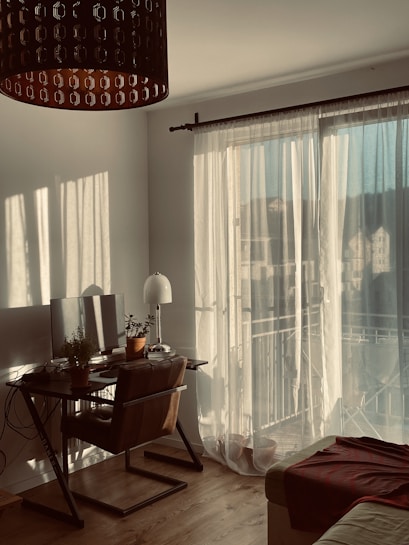 A cozy room filled with warm sunlight filtering through sheer curtains. A wooden desk is adorned with a computer monitor and several potted plants. A comfortable chair is positioned at the desk, and a floor lamp stands nearby. A soft, brown blanket is draped over the edge of a sofa. The wooden floor adds to the inviting atmosphere.