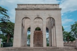 An ornate stone monument featuring three large arches. Above the central arch, the words 'ROMA O MORTE' are inscribed. Behind the arches, trees and a partly cloudy sky are visible, adding a natural backdrop to the architectural structure.