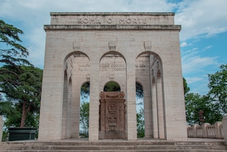 An ornate stone monument featuring three large arches. Above the central arch, the words 'ROMA O MORTE' are inscribed. Behind the arches, trees and a partly cloudy sky are visible, adding a natural backdrop to the architectural structure.