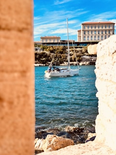 A small sailboat is gently floating on vibrant blue waters, framed by rocky cliffs. In the background, a grand building with classic architecture stands under a bright blue sky with wispy clouds.