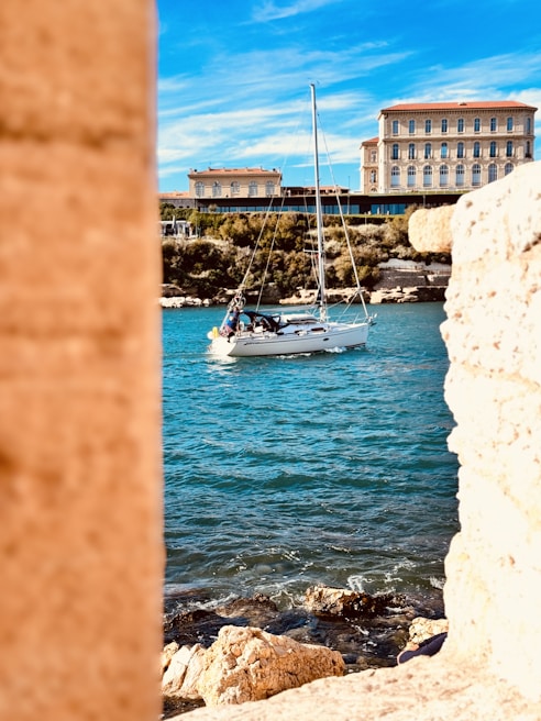 A small sailboat is gently floating on vibrant blue waters, framed by rocky cliffs. In the background, a grand building with classic architecture stands under a bright blue sky with wispy clouds.