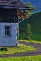 A rustic house with white plaster walls and a dark wooden roof stands adjacent to a winding road. The lush green grass and distant hills suggest a serene countryside setting with a few scattered trees under a clear sky.