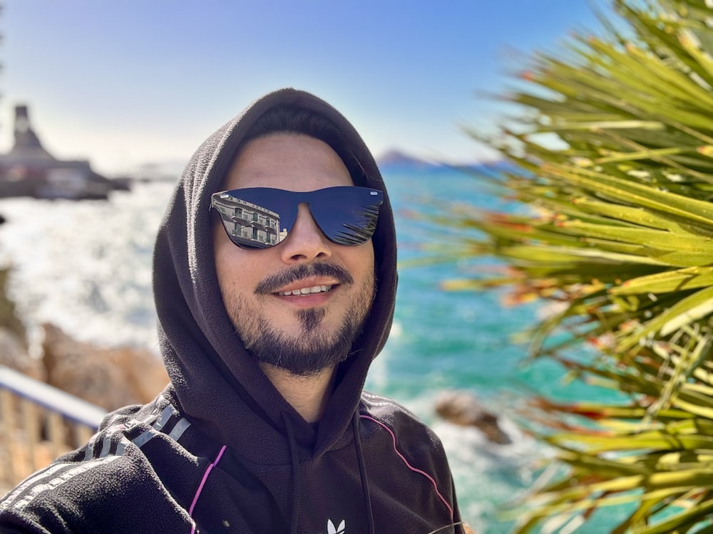 a man wearing a hoodie and sunglasses standing in front of the ocean