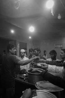 A group of people gathered around a counter in a dimly lit room, reaching out with money or hands towards a man standing behind large pots. The setting appears informal, with visible light bulbs hanging from the ceiling and a slightly chaotic atmosphere.