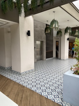 A modern indoor space with decorative tile flooring featuring a blue and white geometric pattern. The area is bordered by neutral-colored walls and pillars. Hanging plants and pendant lights add an element of nature and ambient lighting to the space. Wood accents are visible on the floor and some furniture.