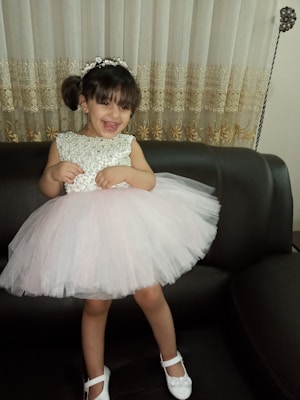 A young girl with dark hair is wearing a white, lace dress with a full tulle skirt. She has a headband with floral embellishments and is smiling widely while standing on a dark sofa. The background features heavily embroidered curtains.