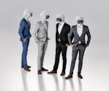 Four figures dressed in suits stand together, each wearing a virtual reality headset and featureless white masks resembling classical sculptures. They appear to be engaged in conversation or interaction, exhibiting a blend of classical and modern elements.