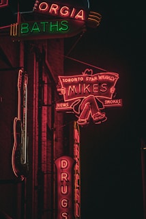 Neon signs illuminate a dark urban scene with vibrant colors. The signs advertise a variety of places and products, with one featuring a figure reading a newspaper. The surrounding architecture is in shadow, contrasting with the bright lights.