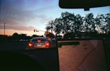 The scene depicts a city street during dusk with a car at a red traffic light. The sky shows a gradient from deep blue to orange, indicating sunset. Palm trees and other foliage are visible on the right, with the silhouette of infrastructure and buildings forming the background. Inside the car, a GPS or navigation device is partially visible, showing a digital map, with blurred focus indicating the arcade's movement.