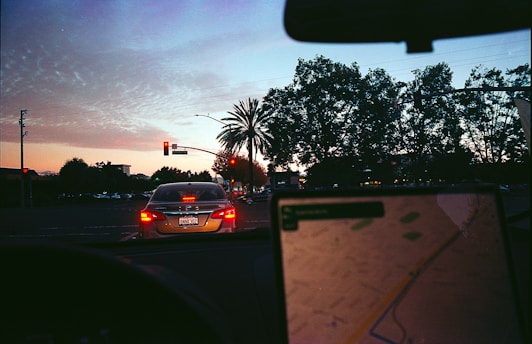 The scene depicts a city street during dusk with a car at a red traffic light. The sky shows a gradient from deep blue to orange, indicating sunset. Palm trees and other foliage are visible on the right, with the silhouette of infrastructure and buildings forming the background. Inside the car, a GPS or navigation device is partially visible, showing a digital map, with blurred focus indicating the arcade's movement.