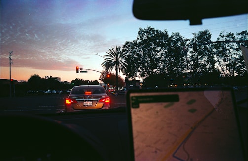 The scene depicts a city street during dusk with a car at a red traffic light. The sky shows a gradient from deep blue to orange, indicating sunset. Palm trees and other foliage are visible on the right, with the silhouette of infrastructure and buildings forming the background. Inside the car, a GPS or navigation device is partially visible, showing a digital map, with blurred focus indicating the arcade's movement.