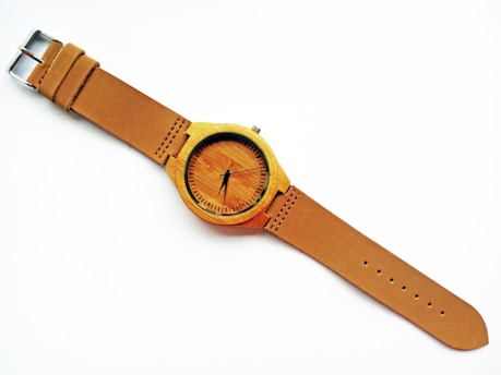 A wooden wristwatch with a tan leather strap and a minimalist design. The watch face has black hour and minute hands with small black lines indicating hours.