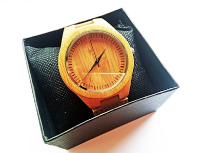 A stylish wooden watch is encased in a black box. The watch features a minimalist design with a wooden face and no numbers, only markers. It has a black hour and minute hand and a wooden strap. The box has a textured black interior, providing contrast with the lighter wood tone of the watch.