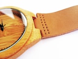 A close-up view of a wooden wristwatch with a smooth, brown leather strap. The watch face is wooden with minimalistic black hour markers and hands. The stitching on the leather strap is visible and neatly aligned.