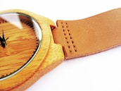 A close-up view of a wooden wristwatch with a smooth, brown leather strap. The watch face is wooden with minimalistic black hour markers and hands. The stitching on the leather strap is visible and neatly aligned.