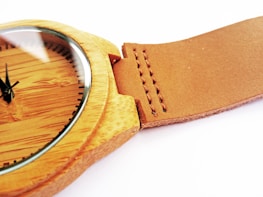 A close-up view of a wooden wristwatch with a smooth, brown leather strap. The watch face is wooden with minimalistic black hour markers and hands. The stitching on the leather strap is visible and neatly aligned.