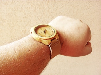 A wooden wristwatch is strapped onto a person's hairy arm. The watch has a natural wood texture and the background is a plain, light-colored surface.